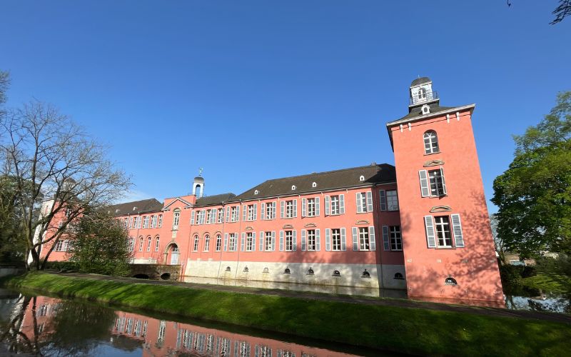 Das Wasserschloss Kalkum mit seiner Rosa Farbe und einem Wassergraben davor.